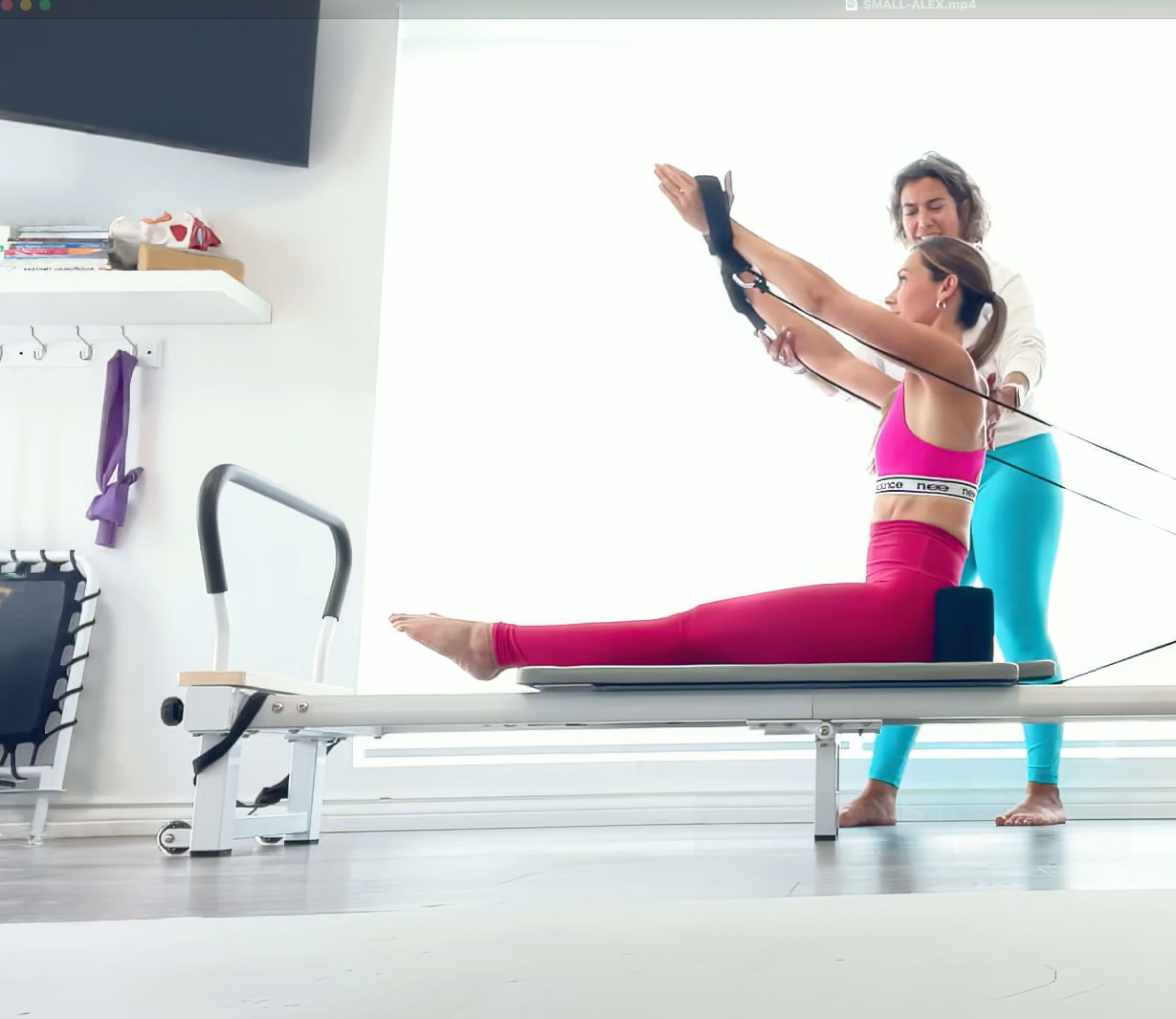 Woman exercising on a Pilates reformer with a trainer in a bright room.